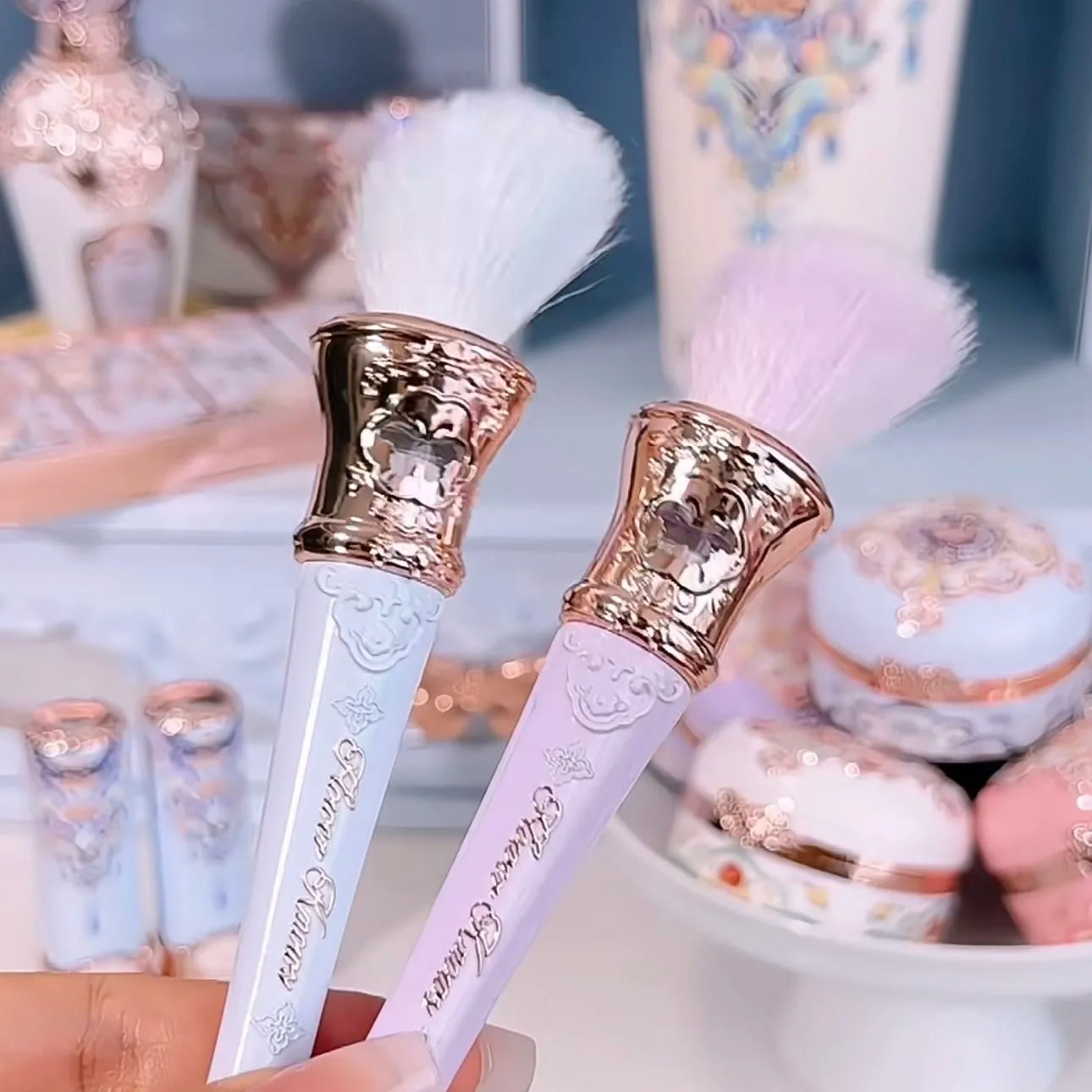 Two decorative makeup brushes with gold accents held by a hand, with a blurred background of donuts and a keyboard.